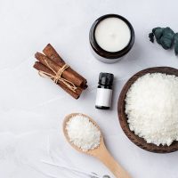 ingredients for candle making , soy wax flakes, candles, cinnamon wicks and wooden spoons on light marble background, blank label for design mockup